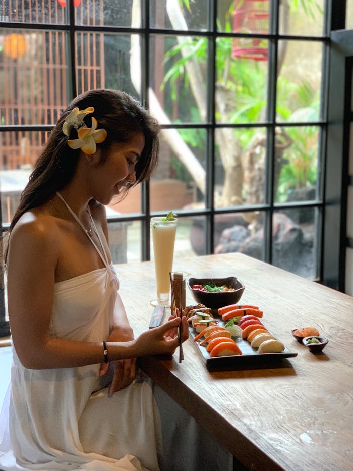 services-img Asian woman savoring a sushi lunch with chopsticks in a cozy indoor setting with a garden view.